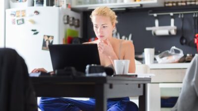 Bord foran vindue med bærbar computer, papir, blyanter, kaffekop og planter.