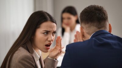 kvinde hvisker til mand, mens en anden kvinde sidder i baggrunden og ser trist ud